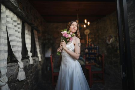 A serene bride holds a pink bouquet, standing in a rustic stone room, capturing her wedding day essence.